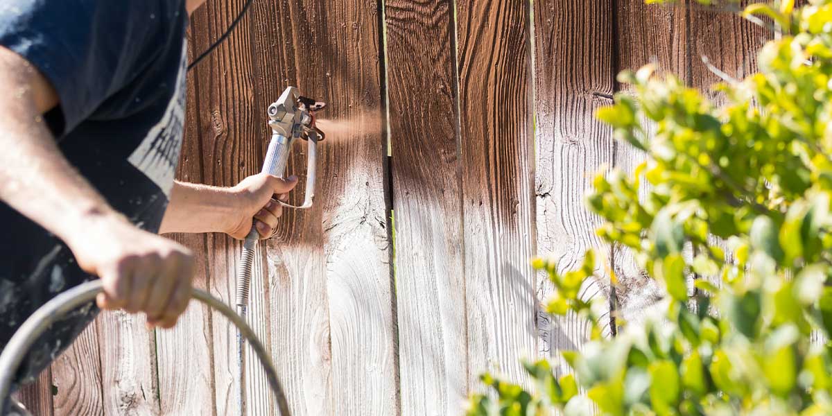 Person spraying a wood fence with stain via a paint sprayer.