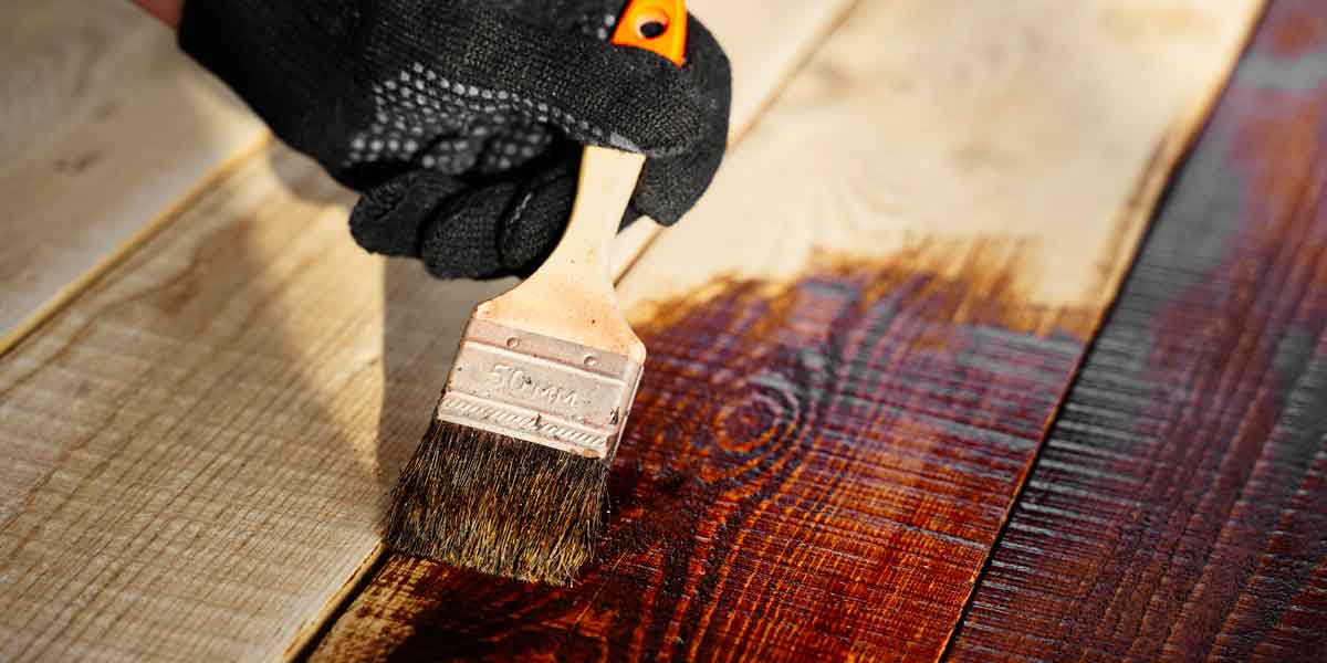 Person brushing on fence stain on a wood panel with a brush while wearing a black glove