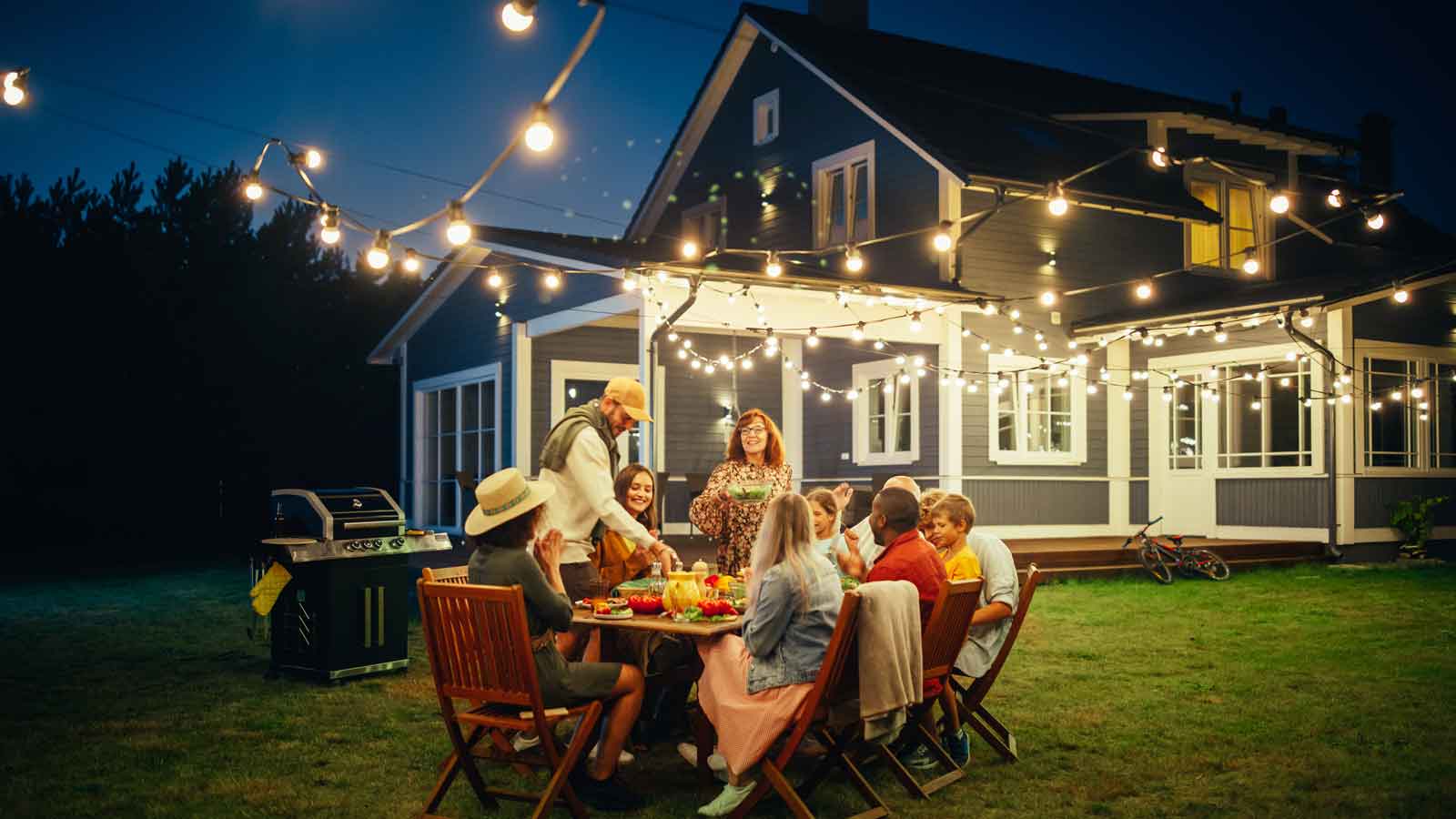 Group of friends and family sit outside during the evening to enjoy a barbeque with string lights overhead.