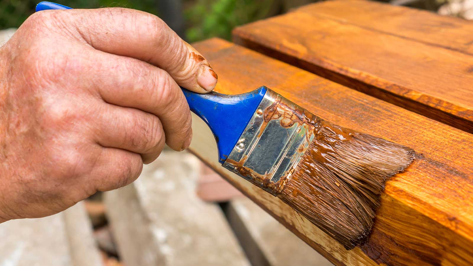 Hand applying a rich, protective wood stain to a deck board using a blue-handled brush, highlighting the hands-on process of high-quality deck staining in Vancouver for longevity.