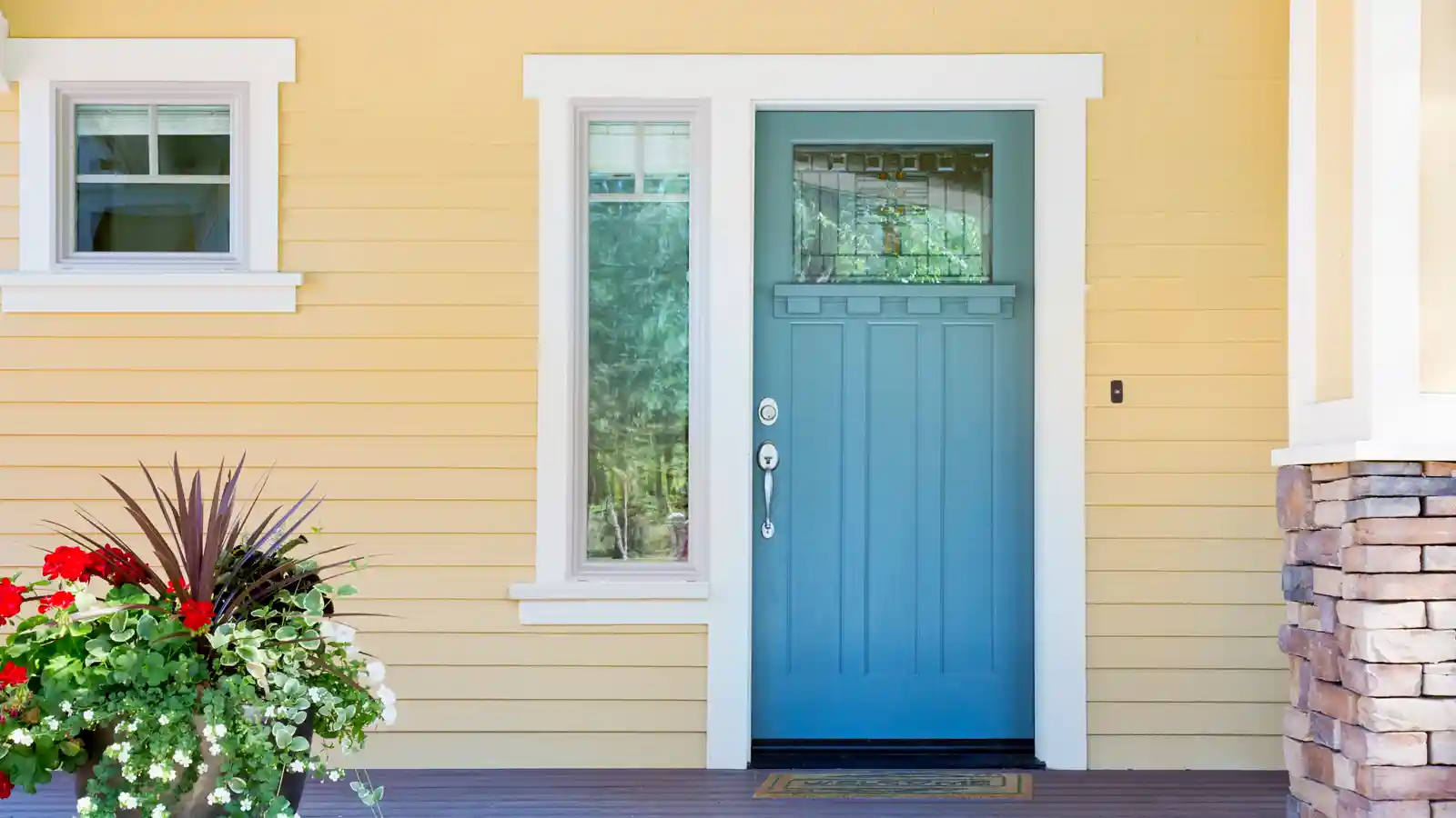 A front entrance of a home with a blue door, yellow siding, and a flowerpot in daytime.